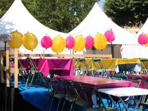 Outdoor party setup with white tents, colorful tables covered in pink and yellow cloths, and folding chairs. Bright yellow and pink lanterns hang above, and trees can be seen in the background.