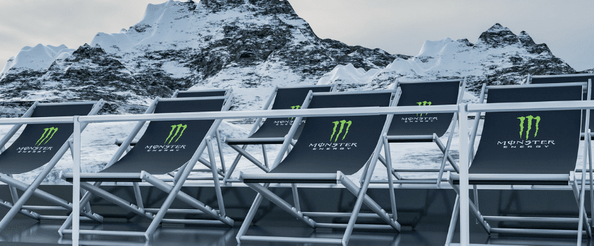 Deck chairs with Monster Energy logos are arranged in a row against a backdrop of snow-covered mountains. The chairs are positioned on a platform with metal railings. The sky is overcast, adding a dramatic feel to the scene.