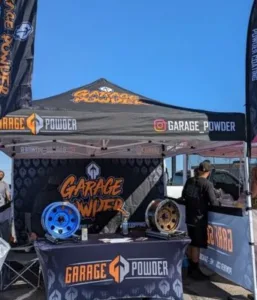 Booth with Garage Powder branding, featuring two alloy wheels on display. The setup includes a branded tent, banners, and a table with promotional materials. A person is standing nearby.