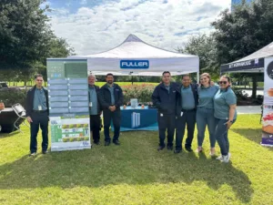 A group of seven people standing in front of a tent with the name Fuller on it. They are outdoors on a grassy area near trees. There is a display board and a table with promotional materials behind them.