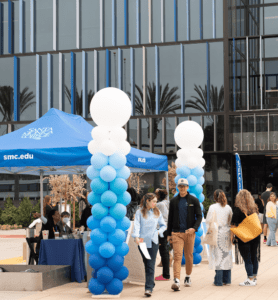 People are gathered outside a building with a Santa Monica College tent. Blue and white balloon columns flank the tent. Some people are walking, while others are standing near a table with information materials. Palm trees are reflected in the windows.