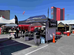 Outdoor event with branded tents displaying products and signs under a clear blue sky. People are exploring the exhibits and chatting. Tall buildings are visible in the background, and orange cones line the area.