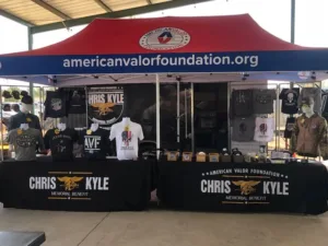 A merchandise booth under a red and blue canopy for the American Valor Foundation. Various shirts and caps featuring military and patriotic designs are displayed on tables, with Chris Kyle Memorial Benefit prominently shown on banners.