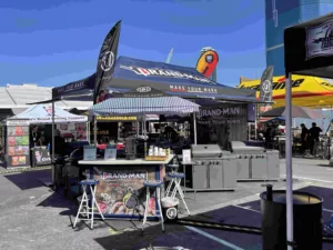 Outdoor event setup with branded tents and signage displaying GRANDMAN: Make Your Mark. Food stalls and counters are visible under the tents. Bright, clear sky and surrounding cityscape in the background.