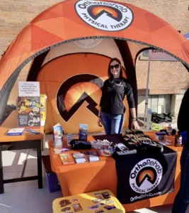 A woman standing under an orange tent with OrthoRehab Physical Therapy branding. The table displays promotional materials, water bottles, and a bowl of candies. The setup is outdoors, in front of a brick wall.