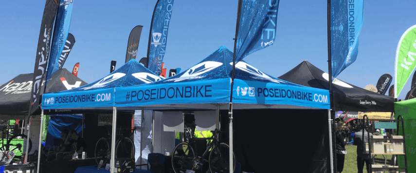 A row of blue tents with Poseidon Bike branding at an outdoor event, surrounded by various flags and banners under a clear blue sky.