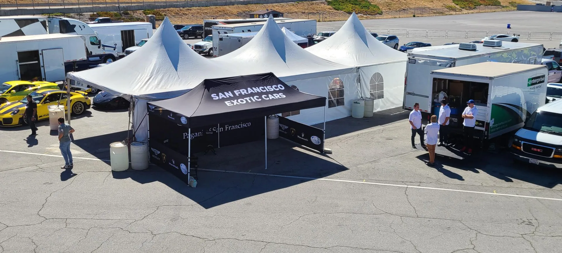 Outdoor setup at a car event featuring a San Francisco Exotic Cars tent alongside multiple white pop-up tents. Parked vehicles and people are around, including a striking yellow sports car.