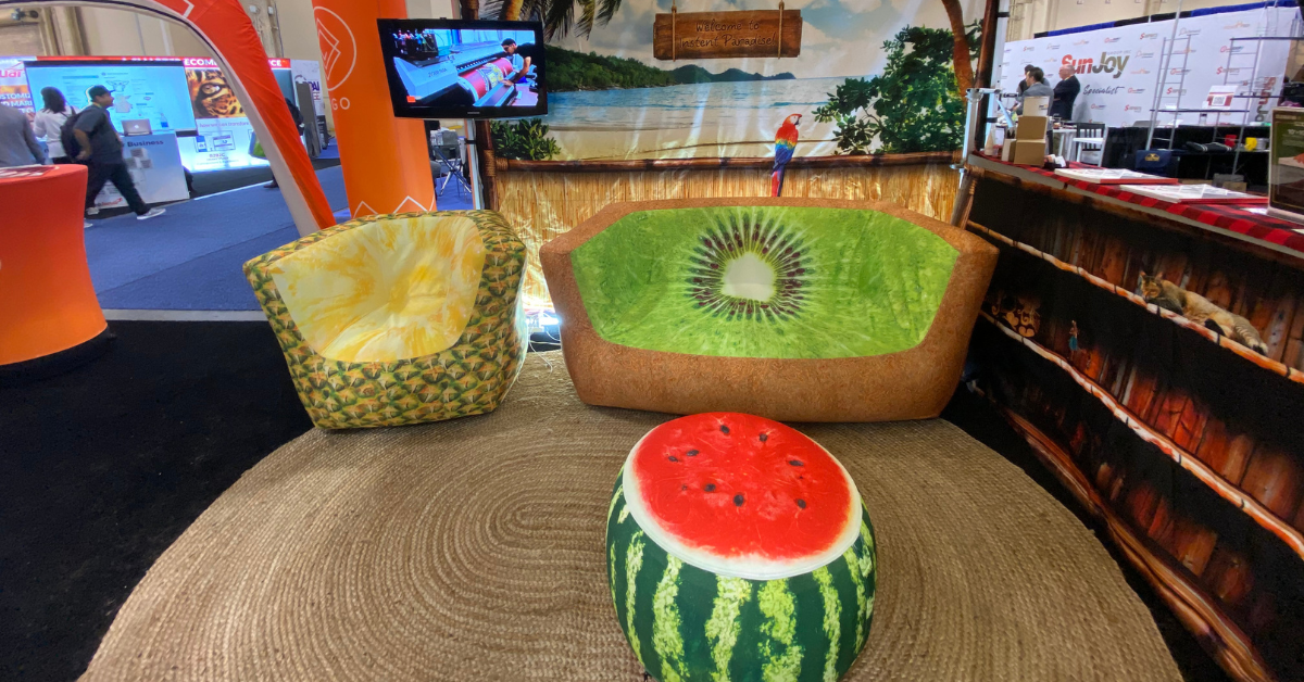 A fruit-themed seating area features chairs designed to resemble a watermelon slice, a pineapple, and a kiwi. Perfect for lively events, a large banner with a beach scene sets the backdrop, while above, a TV screen showcases theme-inspired designs.