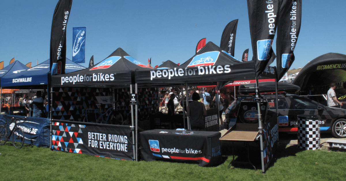 Outdoor bike expo featuring multiple vendor tents, including customized tents from People for Bikes, displaying banners and promotional materials. Attendees are gathered around, enjoying a sunny day.