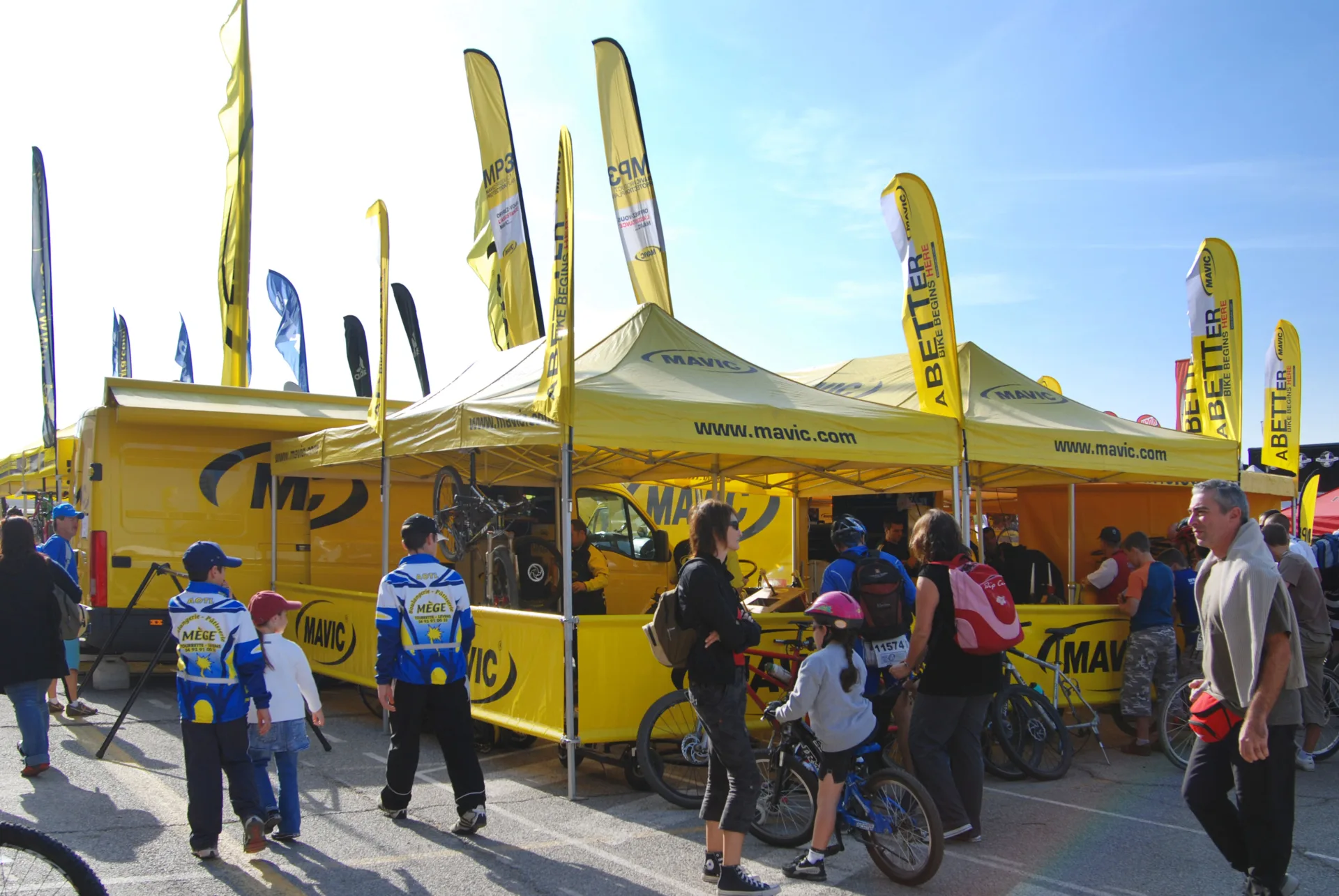 People gather around vibrant yellow Mavic custom tents and vehicles at an outdoor cycling event. Adults and children, some with bicycles, explore the setup under clear blue skies, surrounded by promotional flags.