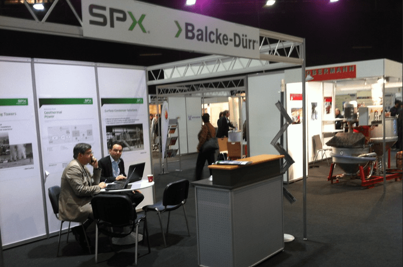 Two men sit and talk at a table in an exhibition booth displaying SPX and Balcke-Dürr signage. The booth features posters and seating. Other booths and attendees are visible in the background.