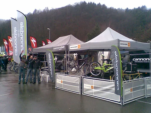 The outdoor bicycle exhibition features custom tents displaying Cannondale bikes. Banners and flags surround the canopies, creating a vibrant scene. Three people are posing in front of the booths with an overcast sky and wooded hills as a picturesque backdrop.