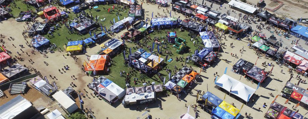 Aerial view of a colorful outdoor event featuring custom tents and canopies arranged in a circular pattern. People stroll along dirt and grass pathways, exploring numerous booths. Vibrant promotional banners and signs adorn the tents, adding to the lively atmosphere.