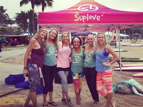 A group of six women smiling under a pink Suplove custom tent. They are on a sandy area with paddleboards nearby, all wearing casual, colorful workout clothes. Palm trees and additional canopies are visible in the background.