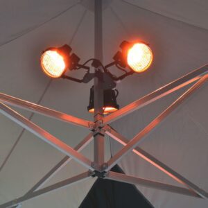 Close-up of the Heating Kit for Pop-Up Canopy, featuring two orange heat lamps mounted on a metallic pole structure inside a tent. The lamps are illuminated, casting a warm glow on the geometric pattern of the tent's ceiling support beams.