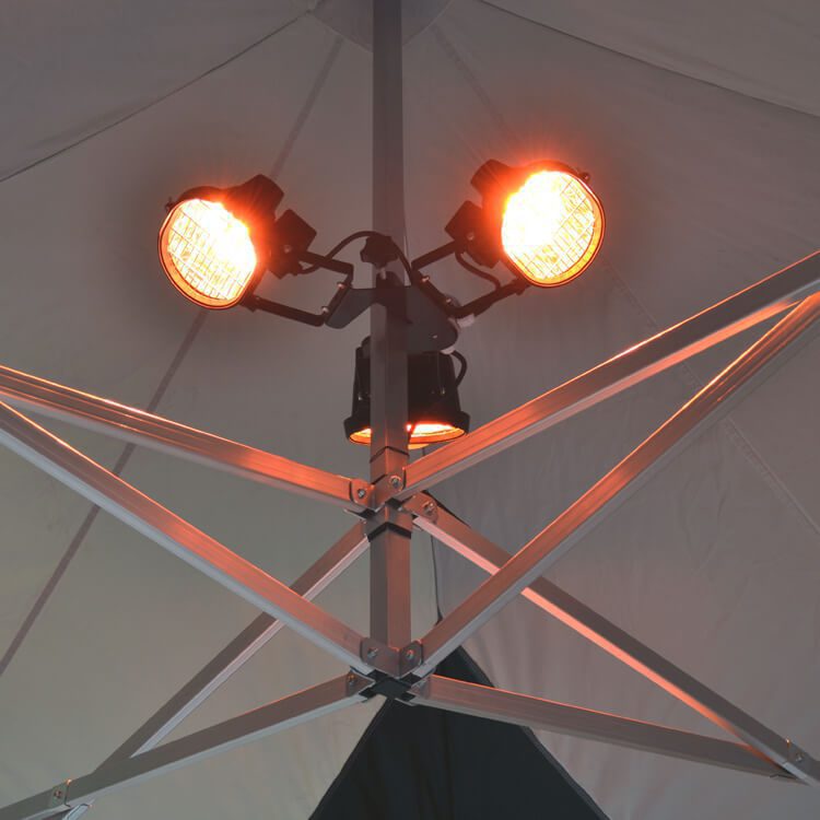 Close-up of the Heating Kit for Pop-Up Canopy, featuring two orange heat lamps mounted on a metallic pole structure inside a tent. The lamps are illuminated, casting a warm glow on the geometric pattern of the tent's ceiling support beams.