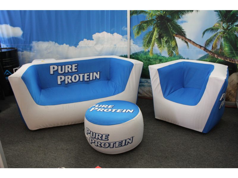 The Custom Air'Lounge Inflatable Furniture Set, in blue and white, includes a sofa, chair, and ottoman all showcasing Pure Protein branding amidst a beach-themed setting with palm trees.