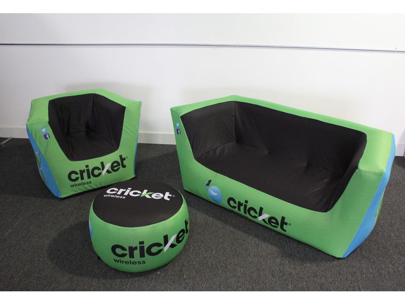 Custom Air'Lounge Inflatable Furniture Set in green and black with Cricket Wireless logos, comprising a loveseat, an armchair, and a round ottoman, arranged on a gray carpeted floor against a white wall.