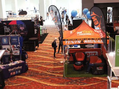 A trade show with various booths and banners displaying brands and products. People walk around the carpeted floor, engaging with exhibitors under custom tents and canopies. Orange and black tents are prominently shown in the foreground.
